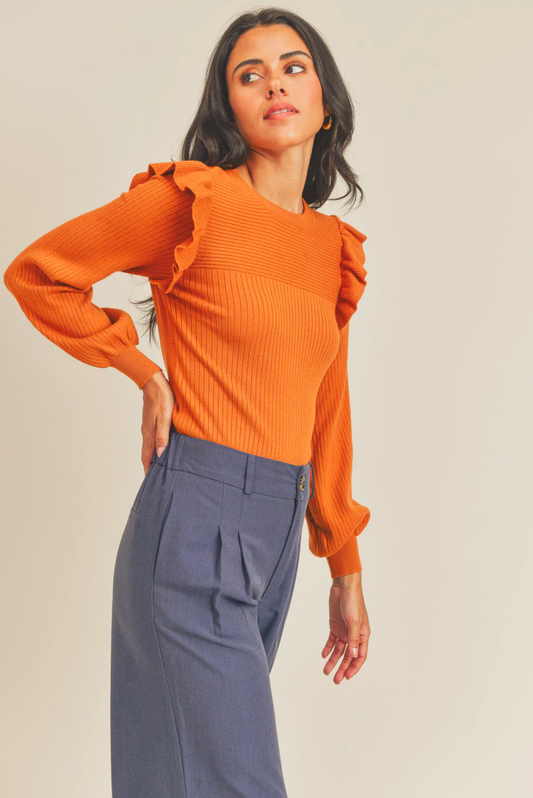 A woman poses in an orange blouse and gray-blue pants.